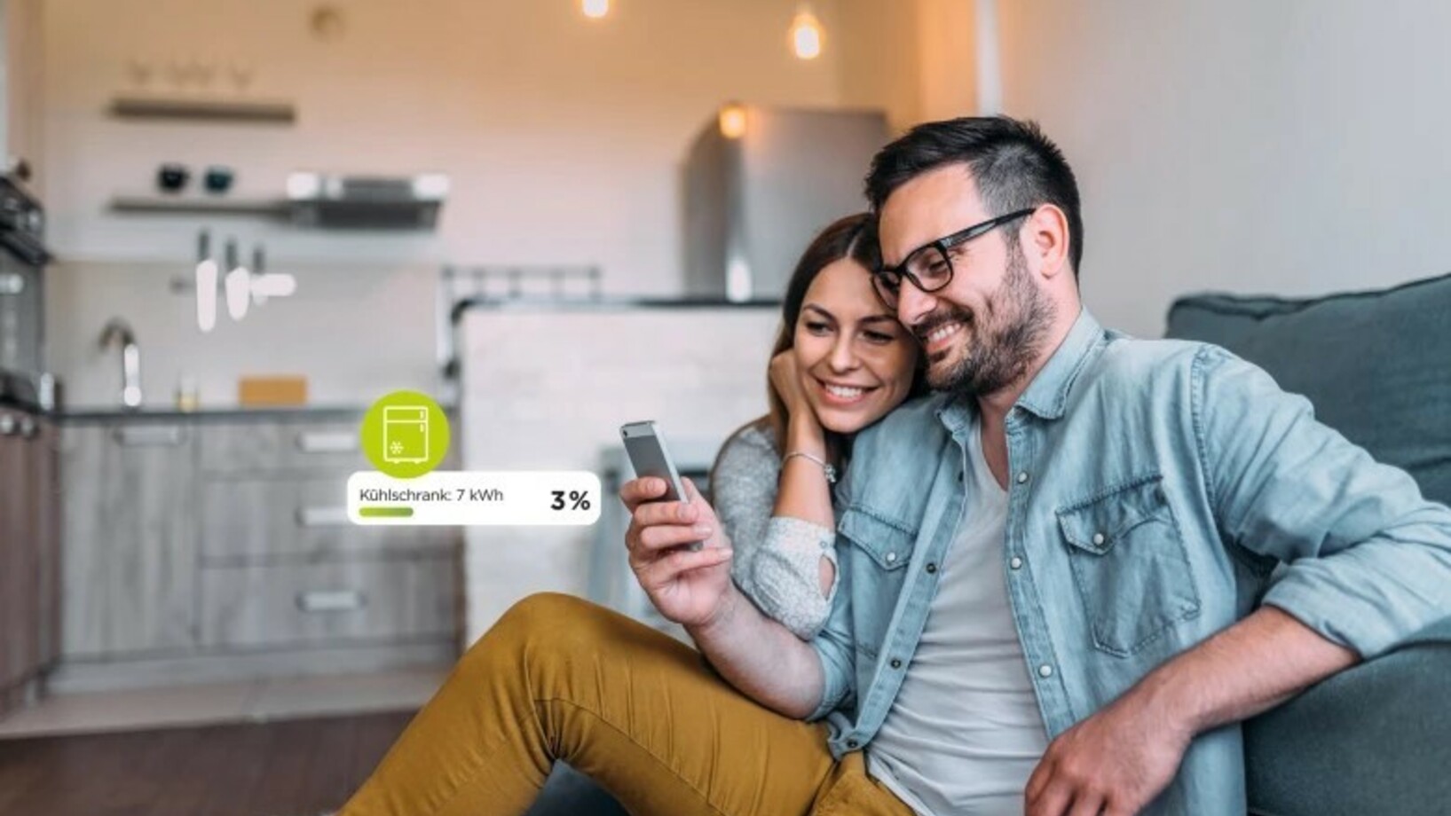 Ein Mann hält ein Handy in der Hand und seine Partnerin schaut an seiner Schulter lehnend mit. Beide lächeln und sitzen angelehnt an eine Couch