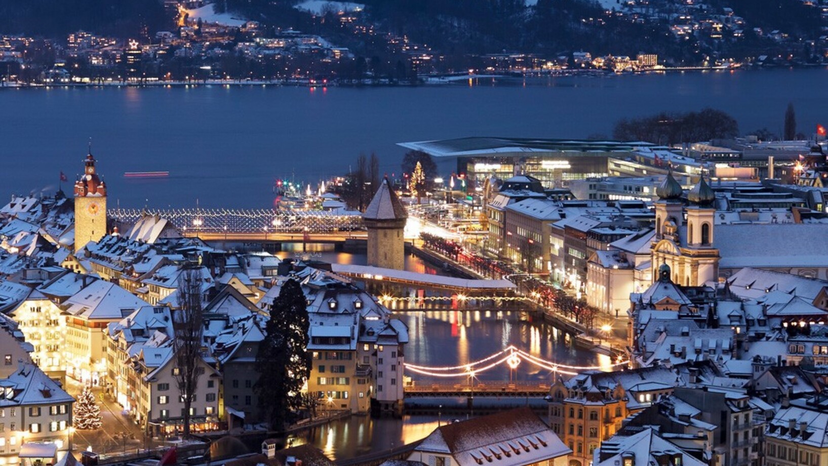 Die Dächer der Stadt Luzern in Weihnachtlicher Beleuchtung mit schneebedeckten Dächern. In der Mitte durchdringt die Reuss die Stadt, im Hintergrund der Vierwaldstädtersee