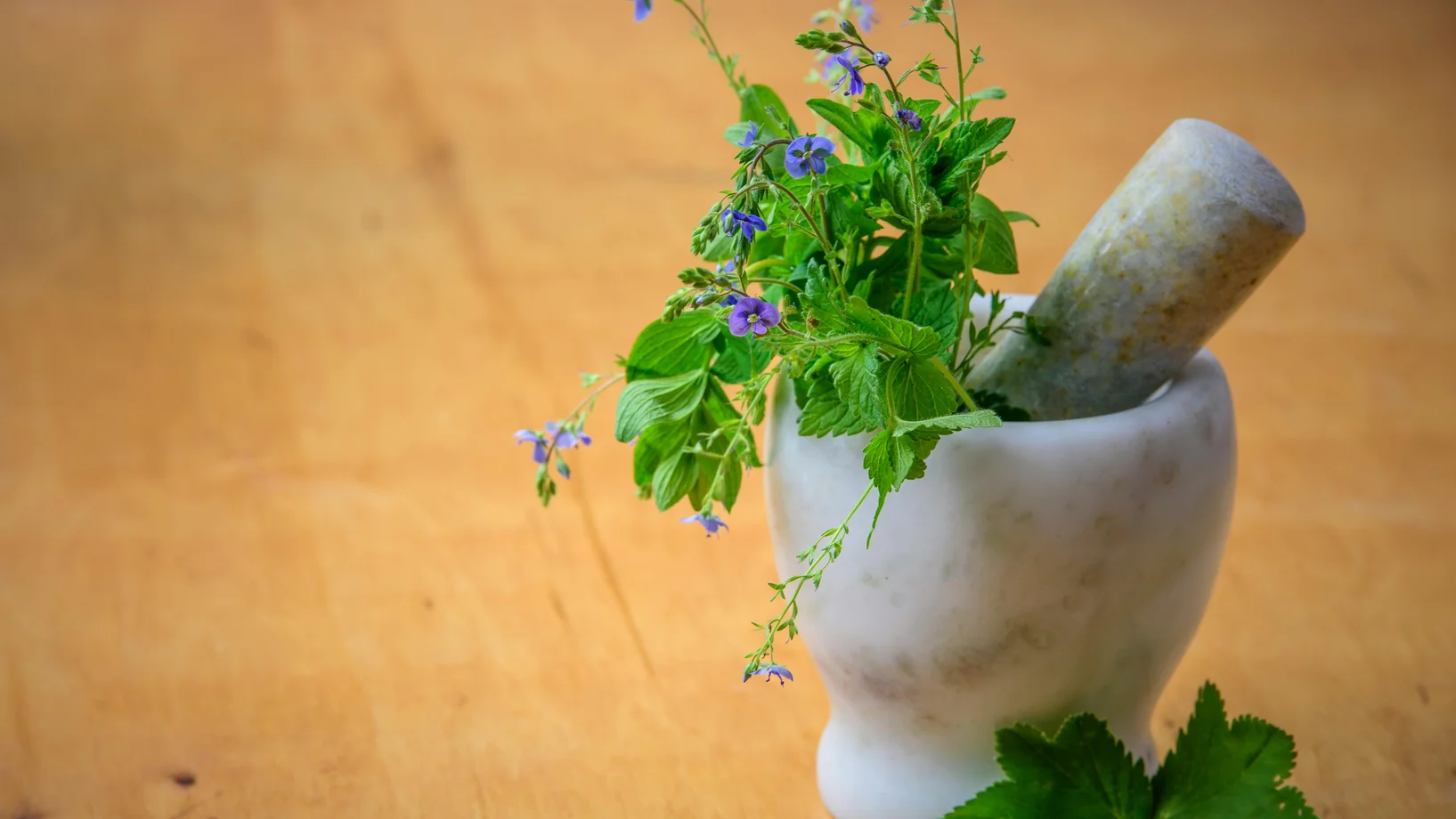 Marble mortar and pestle with fresh herbs and flowers on wooden table enhancing natural health and cooking aesthetics.